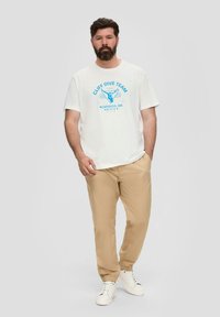 Weißes T-Shirt mit blauem Aufdruck "CLIFF DIVE TEAM", kombiniert mit beigen Hosen mit elastischen Bündchen und weißen Sneakers, vor einem grauen Hintergrund.