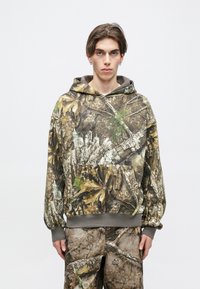 Camouflage hoodie with a drawstring hood, features brown, green, and gray textures resembling bark and leaves. Kangaroo pocket at front.