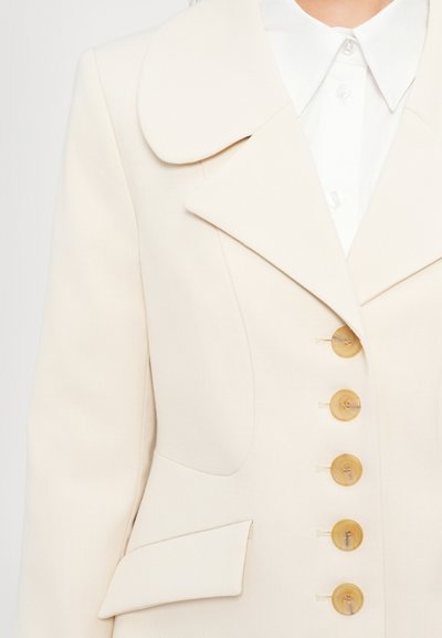 Chaqueta ajustada color beige claro con un gran cuello redondeado, seis botones redondos y un bolsillo lateral. Tela suave con un corte estructurado.