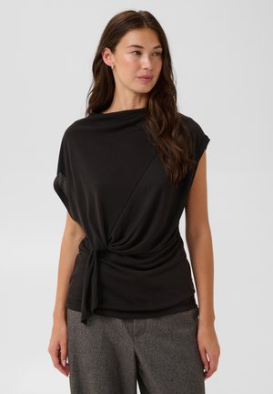 Femme aux longs cheveux bruns portant un haut noir à manches courtes noué à la taille et un pantalon gris, debout devant un fond uni clair.