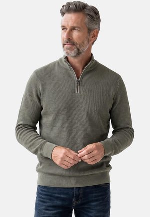 Mann mit grauem Haar und Bart, der einen olivgrünen, strukturierten Viertel-Reißverschluss-Pullover und dunkelblaue Jeans trägt, schaut seitlich mit verschränkten Händen.