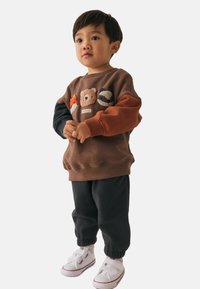 Bambino che indossa una felpa marrone con facce di animali, pantaloni neri e scarpe bianche, in piedi con le mani unite.