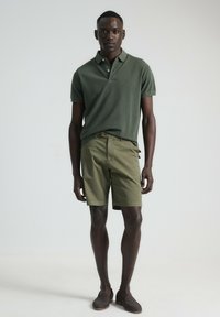 Camiseta polo de color verde oscuro con cuello, pantalones cortos caqui y zapatos de lona trenzados marrones. El modelo está de pie contra un fondo simple.