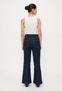 Mulher com cabelo preso em coque, vestindo uma blusa branca sem mangas e calças de ganga escuras de cintura alta e largas, de pé e de costas, em fundo liso.