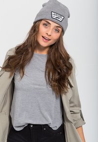Beanie grigia in maglia con patch logo, t-shirt grigia coordinata, giacca beige e pantaloni neri. Il modello ha i capelli mossi e uno sfondo neutro.
