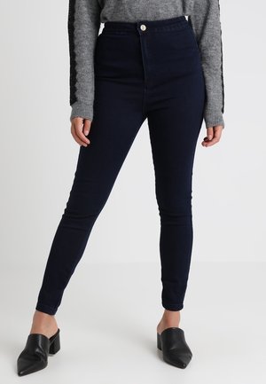 Mujer con jeans ajustados de cintura alta azul oscuro, suéter gris de manga larga y zapatos negros de tacón sin cordones, de pie contra un fondo blanco.