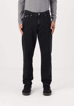 Mann mit schwarzen Jeans im geraden Schnitt, grauem Langarmshirt und schwarzen Sneakers, steht vor einem einfarbigen weißen Hintergrund.