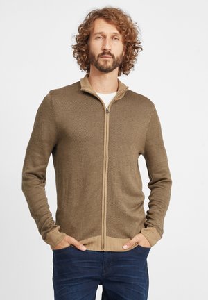 Brun lynlås cardigan lavet af tekstureret strikket stof, med høj krave og ribbede manchetter. Modellen står med hænderne i lommen.