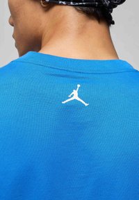 Blaues Baumwoll-T-Shirt mit einem weißen Jumpman-Logo auf dem oberen Rücken. Der Stoff hat eine glatte Textur und ein klassisches Rundhalsausschnitt-Design.