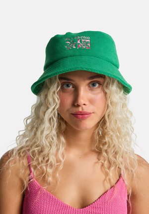 Groene emmerhoed van zacht materiaal met geborduurd bloemenpatroon en merknaam, voorzien van een brede, losse rand voor bescherming tegen de zon.