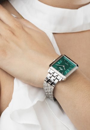 Orologio quadrato con quadrante verde, cinturino in acciaio inossidabile argentato e indici delle ore luminosi. Indossato su un polso coperto da un tessuto bianco.