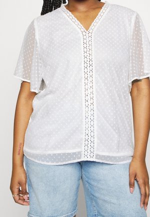 Person iført en hvid semi-gennemsigtig bluse med flagrende ærmer og blondekant, kombineret med lyseblå denimshorts.