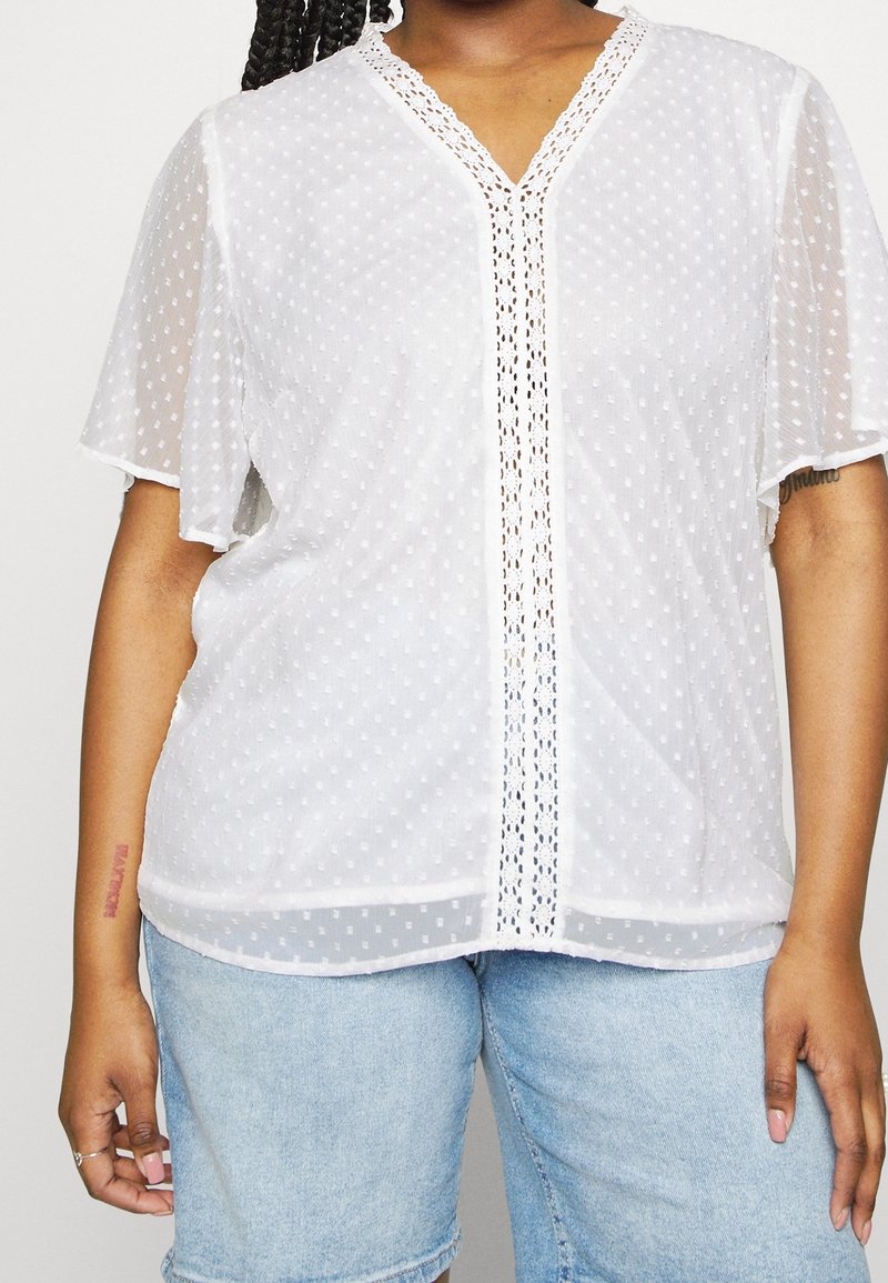 Personne portant un chemisier blanc semi-transparent avec des manches volantées et une bordure en dentelle, associé à un short en jean bleu clair.