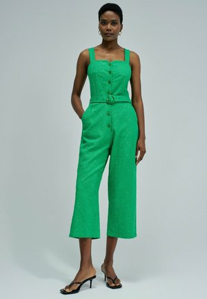 Femme en combinaison verte sans manches avec ceinture, boutons sur le devant et pantalon large court, portant des sandales noires à talons ouvertes, debout les mains dans les poches.