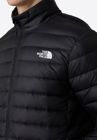 Giacca nera isolata con cuciture orizzontali, colletto alto e chiusura con zip. Presenta un logo bianco sulla zona sinistra del petto.