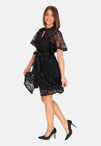 Robe en dentelle noire avec des manches courtes et évasées, un devant à boutons et une ceinture à la taille. L'ourlet a un bord festonné, offrant une finition délicate.