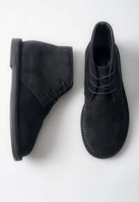 Czarne zamszowe botki za kostkę o zaokrąglonym kształcie, z ozdobnymi sznurowadłami, gładką fakturą i płaską gumową podeszwą. Projekt z minimalną ilością przeszyć.