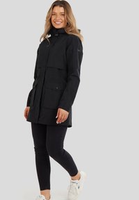 Chaqueta negra impermeable con un cuello alto, bolsillos delanteros y cierre de botón, llevada sobre jeans oscuros y zapatillas blancas.