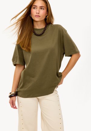 T-shirt olive vert à manches courtes en tissu doux, associé à un pantalon large crème avec des accents dorés le long des coutures latérales.