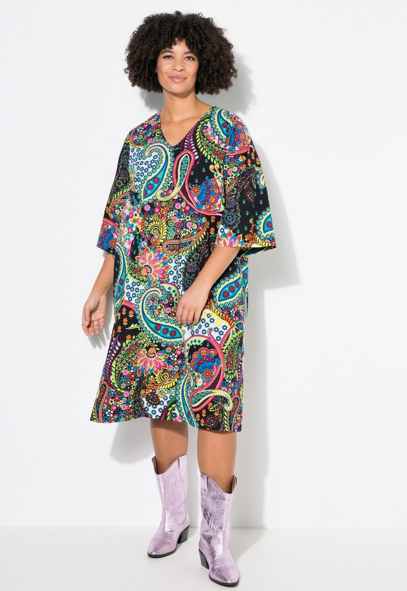 Vestido de estampado paisley colorido con escote en V y mangas anchas, hecho de una tela ligera. Combinado con botas de vaquero brillantes en color lila.