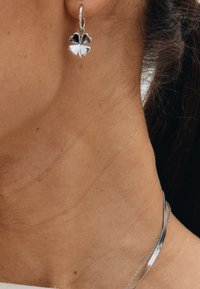 Sterlinghõbedast rõngaskõrvarõngas, millel on neljaleheline ristikujundus vahelduvate hõbedaste ja mustade aktsentidega. Kanda siledal, päevitunud kaelal.