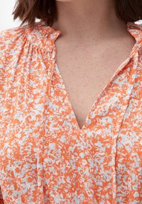 Bluzka w pomarańczowo-białe kwiatowe wzory z dekoltem w kształcie litery V, zebranymi ramionami i lekkim materiałem. Charakteryzuje się delikatną teksturą i miękkim układem.
