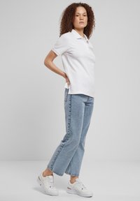 Bijela polo majica s kratkim rukavima, svijetlo plave traperice ravnog kroja i bijele tenisice. Model stoji opušteno, s jednom rukom na bokovima.