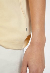 Żółta bawełniana koszulka z rozcięciem po bokach, zaokrąglonym dołem i małą metką "Guess" na dolnym brzegu, w zestawieniu z białymi spodniami i widoczną ręką.