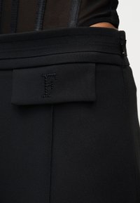 Κοντινή λήψη μαύρου ενδύματος ψηλής μέσης με ραμμένο γράμμα «F» σε τσέπη με καπάκι, φορεμένο πάνω από μαύρο ριμπ μπλουζάκι.