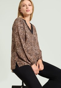 Blusa de estampado de leopardo en marrón claro con escote de encaje negro, mangas largas y aberturas laterales. Combinada con pantalones ajustados negros.