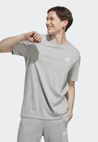 Camiseta de algodón gris con mangas cortas, cuello redondo y logo blanco de Adidas en el lado izquierdo del pecho. Textura suave y corte relajado.