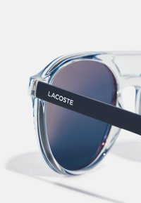 Lacoste napszemüveg átlátszó kerettel és sötét gradiens lencsékkel. Kék díszítésű vékony szárak fehér "LACOSTE" felirattal.