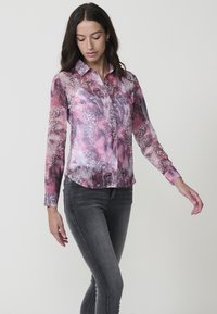 Blusa transparente con un estampado abstracto de animal en tonos rosa, púrpura y gris, con cuello de camisa y mangas largas, combinada con jeans ajustados oscuros.