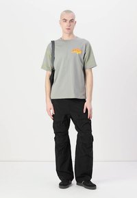 Grå bomullst-skjorte med oransje grafikk på brystet, paret med svarte cargo-bukser med flere lommer og en avslappet passform. Svarte joggesko.