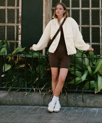 Bela flis jopica preko rjave bodi obleke, v kombinaciji z belimi supergami in gležnjarji. Stoji ob zelenju s hrbtno stranjo opečne poti.