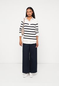 Gestreifter Pullover in Weiß und Marineblau, weite dunkle Jeanshose und weiße Sneakers. Trägt eine kleine beiges Umhängetasche.