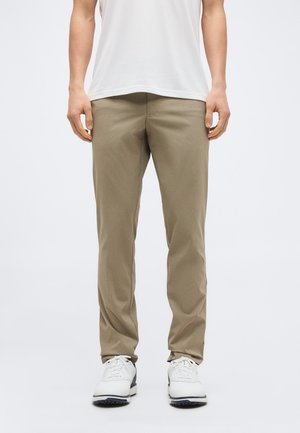 Mand iført beige bukser med lige pasform, hvide sneakers og en hvid t-shirt, stående mod en enkel lys baggrund.