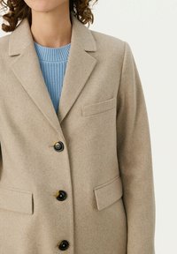 Beige Wollsakko mit Reverskragen, einer einzelnen Brusttasche und schwarzen Knöpfen. Über einem hellblauen Strickpullover getragen.