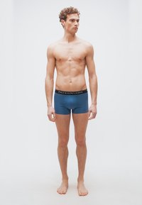 Νεαρός άνδρας με σγουρά μαλλιά στέκεται ξυπόλυτος φορώντας μπλε εσώρουχα Polo Ralph Lauren μπροστά σε απλό λευκό φόντο.