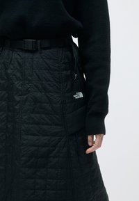 Saia preta acolchoada com um tecido texturizado, cinto ajustável e bolsos laterais. Detalhe do logo visível no lado inferior direito.