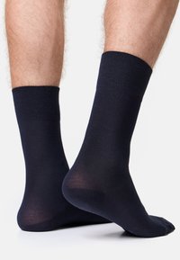 Calcetines azul marino cubren tobillos desnudos, resaltando una textura suave y un ajuste ceñido. Los pies están colocados uno al lado del otro sobre un fondo liso.