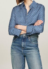 Sininen nappipaidan paita, jossa on geometrinen valkoinen kuvio, pitkät hihat käärityt; yhdistetty korkeavyötäröisiin sinisiin farkkuihin.