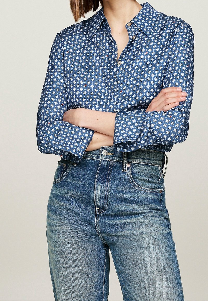 Sininen nappipaidan paita, jossa on geometrinen valkoinen kuvio, pitkät hihat käärityt; yhdistetty korkeavyötäröisiin sinisiin farkkuihin.