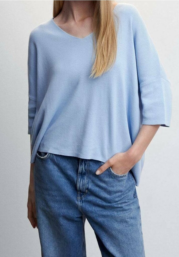 Pull bleu clair léger à col en V avec manches trois-quarts, porté avec un jean bleu taille haute, une main dans la poche, sur un fond neutre.