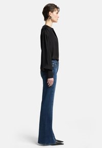 Zwarte satijnen blouse met puffed mouwen, gecombineerd met uitlopende blauwe spijkerbroek. Zwarte puntige schoenen accentueren de outfit. Zijaanzicht.