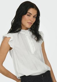 Blusa blanca sin mangas con hombros de volantes, un frente abotonado y una tela texturizada, que presenta líneas verticales sutiles y un corte holgado.