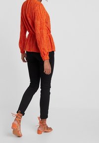 Orange strukturierte Bluse mit langen Ärmeln und tailliertem Schnitt, kombiniert mit schwarzen Slim-Fit-Jeans und orangefarbenen Riemchen-Heels.
