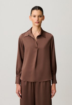 Femme aux cheveux foncés coiffés en chignon bas, portant un chemisier en satin brun ample à manches longues et col large, sur fond clair uni.