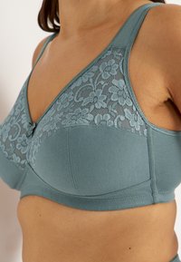 Nahaufnahme einer Person, die einen blaugrünen BH mit floralen Spitzenverzierungen auf den Cups und breiten Schulterträgern trägt.