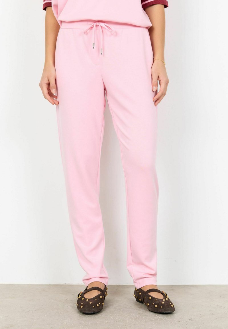 Ružové tepláky s nastaviteľným pásom na šnúrku, vyrobené z mäkkej látky, s úzkym strihom a bočnými vreckami. Doplnkom sú tmavohnedé topánky so zlatými detailmi.